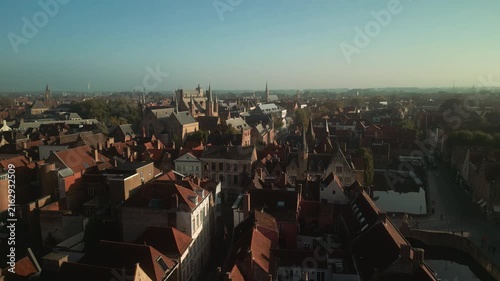 Elevated drone view across Bruges, Belgium, reveals dense medieval rooftops, narrow streets and winding waterways extending toward the hazy historic skyline.