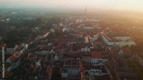 Warm sunrise over Bruges, Belgium, reveals dense red rooftops, historic houses and distant church spires across the atmospheric medieval cityscape at dawn.