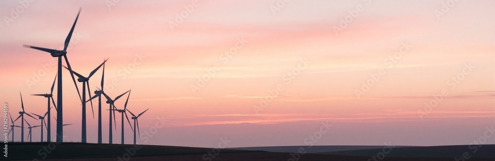Fototapeta premium Wind turbines stand silhouetted against a soft pastel sunset sky in a rural field. The farm generates clean energy at twilight, with rotors slowly turning and blades visible against the horizon.