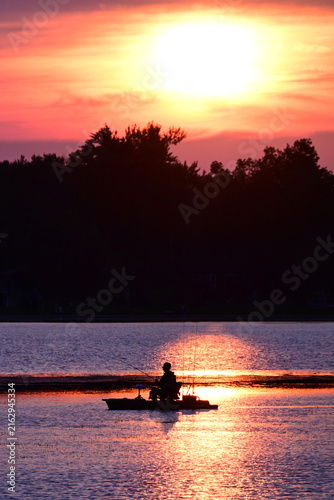 A lone fishermen sitting on a platform boat fishing boat as the sun sets behind him