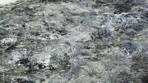 Water flows over rough stones. Tiny bubbles form as current moves fast. Surface reflects light in shimmering patterns. Ideal for nature, adventure, or eco-themed projects. Can be used in travel