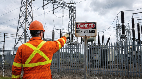back view of electrician in safety suit pointing to danger high voltage sign at power substation industrial electricity grid worker high risk energy facility professional utility maintenance safety pr