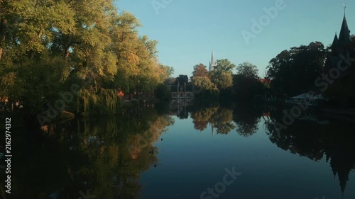 Waterfowl cross a quiet lake in Bruges, Belgium, surrounded by autumn trees and deep reflections, while distant historic buildings catch the warm light of dawn.