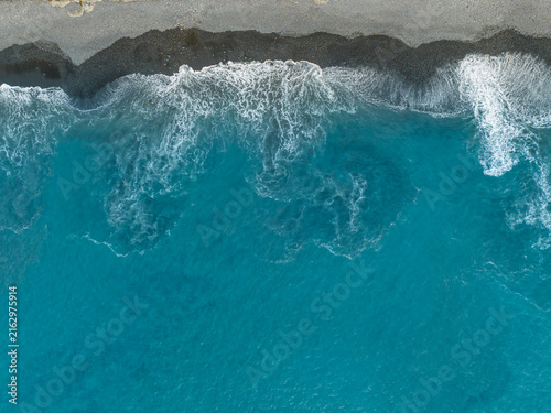 Aerial view of turquoise ocean waves crashing against the dark volcanic sand beach in Madeira, Portugal.