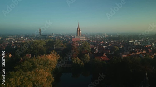 Aerial panorama across a tree-lined lake reveals the historic skyline of Bruges, Belgium, with prominent church towers, red roofs and autumn foliage at dawn.