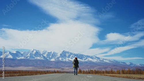 Lone traveler walks down a straight road toward distant snow-capped mountains. Blue sky with wispy clouds stretches overhead. Person carries backpack, suggesting adventure or exploration