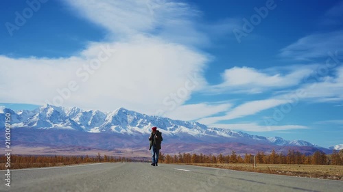 Lone traveler walks down a wide open road toward distant snow-capped mountains. The sky is vast and blue with wispy clouds. Autumn foliage lines the roadside. The person appears calm, focused