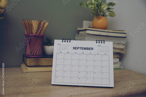 2026 Calendar on wooden desk in indoor workspace with books, pencil holder and flowers for monthly planning, scheduling and organization.