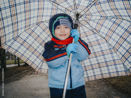 Fotografie Young child wearing blue winter jacket and red scarf holds a plaid umbrella whil