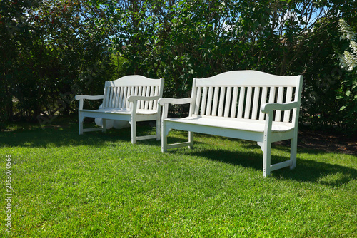 Elegant white wooden benches standing on green grassy lawn in summer garden. Luxury backyard relaxation zone surrounded by high green bushes and blue sky. Beautiful outdoor seating area for romantic.