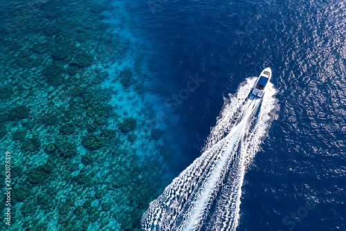 Aerial view of luxury yacht speeding through deep blue ocean water near coral reef.
