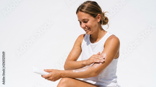 Young woman applying sunscreen lotion to her arm with a tube on a bright white background