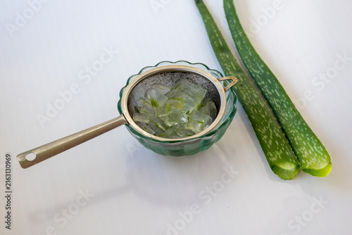 Fotografía Fresh diced aloe vera gel cubes inside metal strainer over glass bowl with green leaves on white background