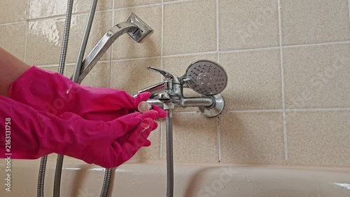 A hand in a vivid pink glove works on the chrome shower mixer and showerhead in a tiled bathroom; the close-up captures the precise interaction with the plumbing fixture during cleaning