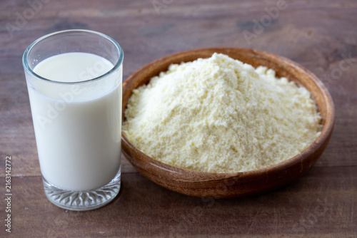 Fresh liquid milk filled in transparent glass served beside dry powdered milk in wooden bowl. Natural dairy nutrition product on wooden table background.