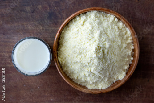 Transparent glass filled with fresh milk placed beside dry powdered milk in wooden bowl. Healthy dairy ingredient served on rustic wooden tabletop surface. Top view.