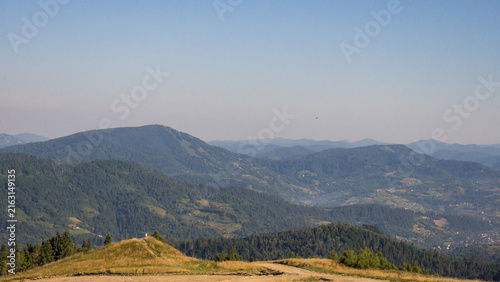 Konstfotografi Aerial landscape photograph of a vast mountain valley filled with dense pine forests, scattered rural houses, and light morning mist under layered blue ridges