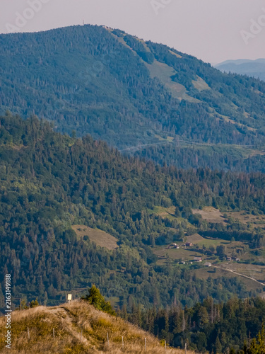 Konstfotografi Aerial landscape photograph of a vast mountain valley filled with dense pine forests, scattered rural houses, and light morning mist under layered blue ridges