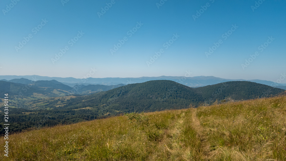 Fototapeta premium High-angle panoramic view of rolling mountain ridges, sloping grassy hillside, and expansive clear blue sky on a sunny day.