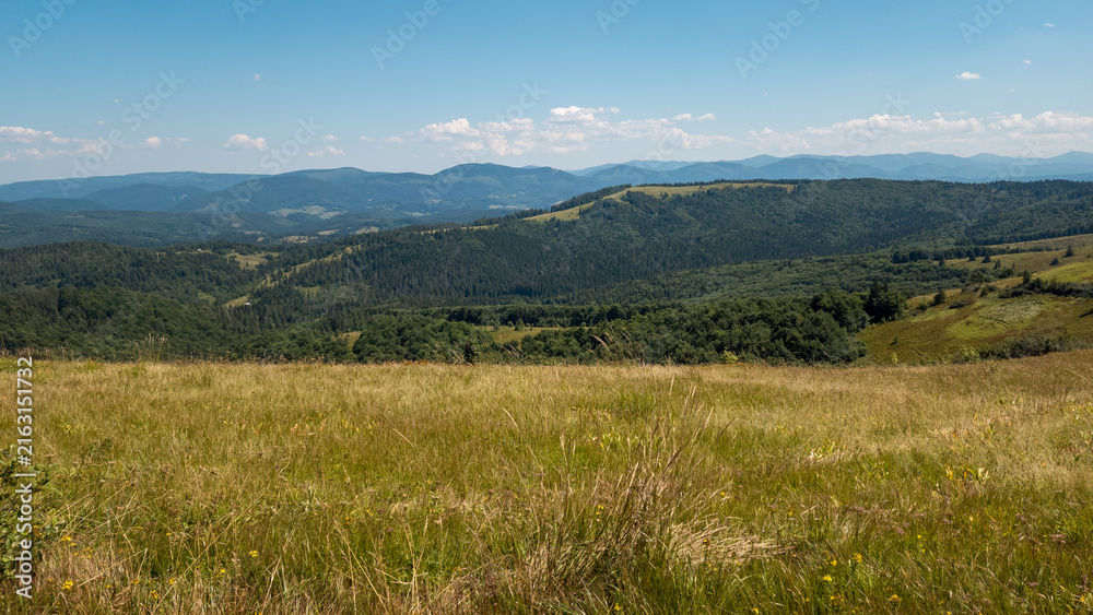 Fototapeta premium Wide outdoor stock photo of a narrow dirt trail stretching across a grassy alpine mountain ridge toward distant blue peaks under a vast clear blue sky. Concept for trekking journey, eco adventure, tra