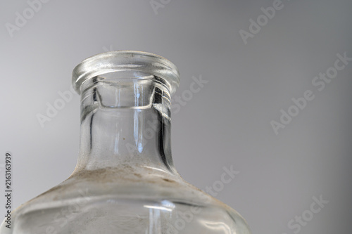 Close-up of a clear glass bottle neck positioned on the left side. Shows highlights, reflections, and dust particles against a gradient gray background.