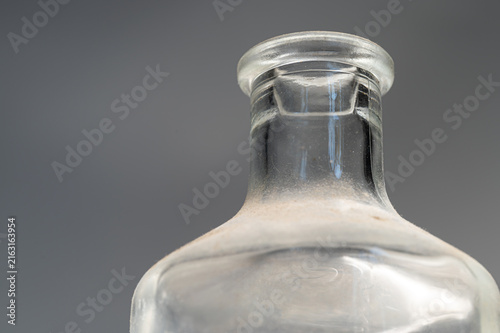 Macro shot of a clear glass bottle neck against a solid gray background. Highlights and reflections are visible on the curved transparent surface.