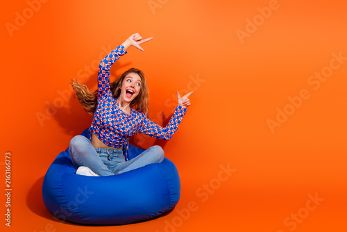 Excited Young Woman in a Stylish Checkered Pullover Posing on a Bean Bag with Bright Orange Background