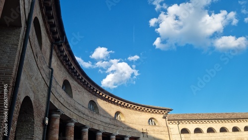 Historic curved brick architecture with arched windows beneath a vivid blue sky and white clouds, viewed from a courtyard.