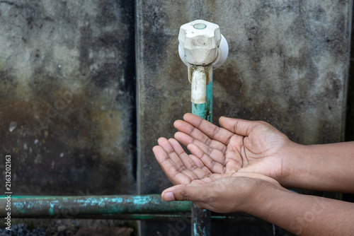 Cupped hands waiting for drop under dirty plastic water tap against concrete wall. Powerful visual for water crisis, climate change, and hygiene issues.