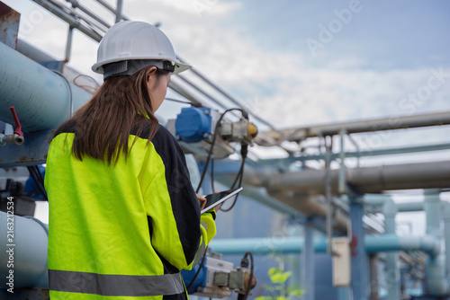 Female engineer inspecting water treatment pipelines with a digital tablet at a water supply plant. Concept of infrastructure, utility maintenance, water management and safety.