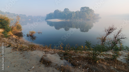A serene, misty morning landscape captures a calm river reflecting a small, tree-covered island under a soft, hazy sky