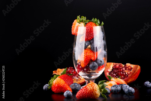 Studio shot of a crystal glass with fresh red berries inside and around it, set against a black background, positioned in the corner.