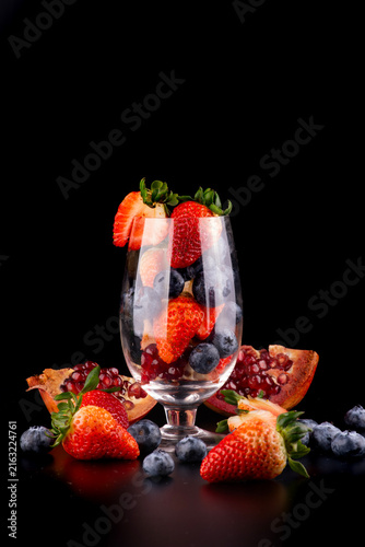 Crystal glass with red fruits—strawberry, pomegranate, and blueberry—inside and around it, against a black background; studio shot.