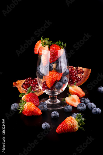 Studio shot of a glass with red fruits—strawberries, pomegranate, and blueberries—inside and around the base; a healthy, black-background image.