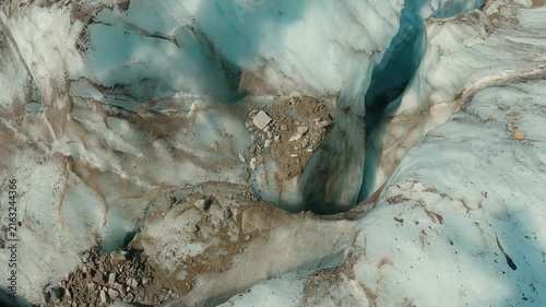 Aerial view of glacier surface showing icy textures, scattered rocks, deep blue meltwater channels. Cool tones dominate with white, gray, turquoise hues