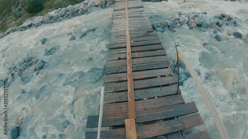 Wooden suspension bridge spans rushing river with rocky banks. Water is milky and turbulent. First person perspective looking down at planks. Adventure travel concept