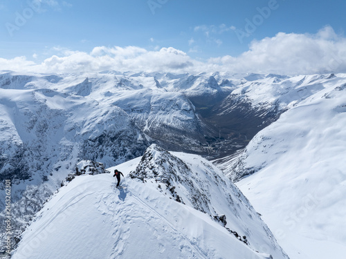 Aerial view of a skier traversing the snowy ridge of Ringshornet with vast mountain peaks and deep valleys under a clear blue sky in Fjord, More og Romsdal, Norway.