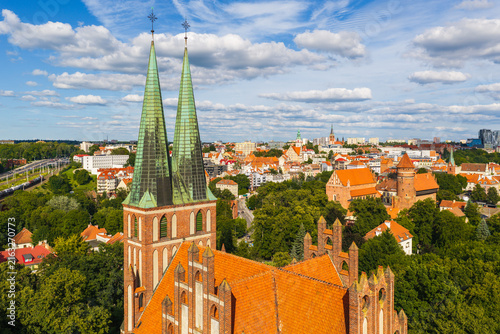 Aerial view of the Garrison Church in Olsztyn with twin spires rising above red-tiled rooftops and lush green trees in the historic city center of Olsztyn, Warmian-Masurian Voivodeship, Poland.