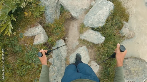 Hiker in blue pants green shirt uses trekking poles to navigate rocky mountain path. Green grass wildflowers surround trail. Natural daylight highlights rugged terrain active outdoor adventure mood