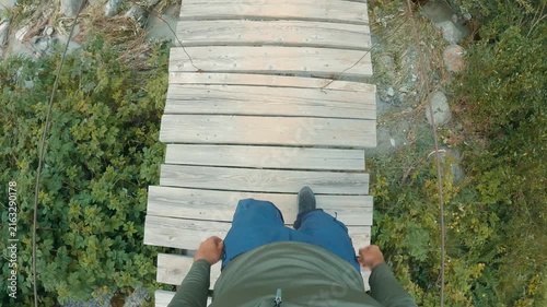 A first-person view of a tourist walking across a dilapidated wooden suspension bridge over a turbulent rocky stream. Natural daylight evokes associations with adventure and nature exploration