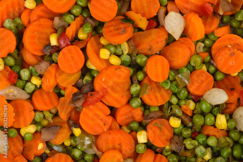 A top down full frame view of a colorful mix of cooked sliced carrots green peas sweet corn kernels and pieces of mushrooms.