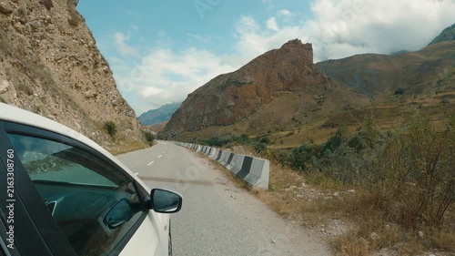 Winding asphalt road through rugged mountain landscape with rocky cliffs green valley. White car in foreground, concrete barriers along roadside. Bright daylight, blue sky with scattered clouds