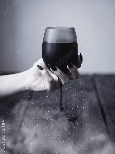 Hand with black manicure holding dark wine glass over rustic wooden table, dust particles, moody gothic still life.