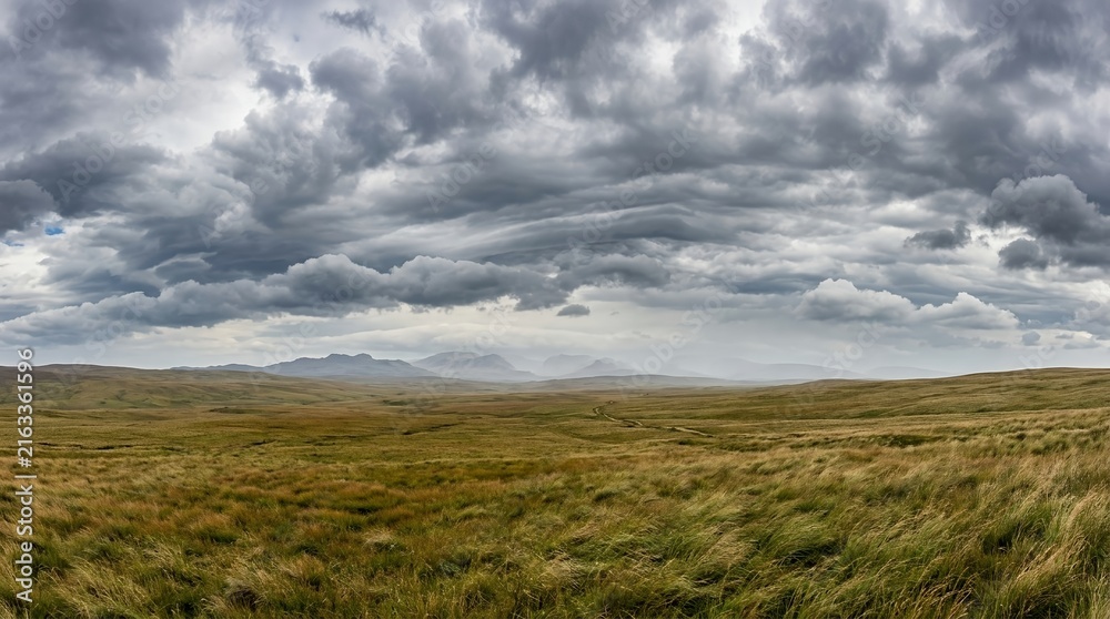 Fototapeta premium Dramatic Stormy Sky Over Vast Grassy Moorland Landscape with Distant Mountain Peaks and Moody Overcast Atmosphere