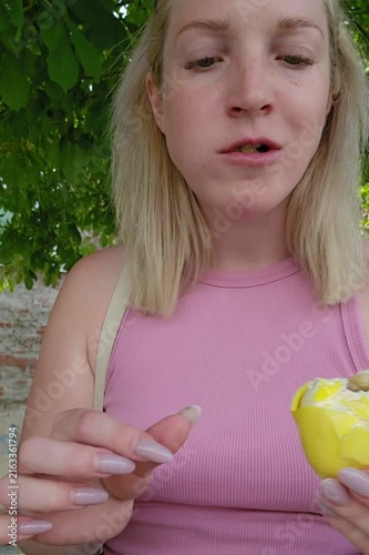 A woman with blonde hair displays a partially bitten lemon-shaped molded shell pastry; her puckered expression highlights the tangy flavor profile, framed by verdant greenery