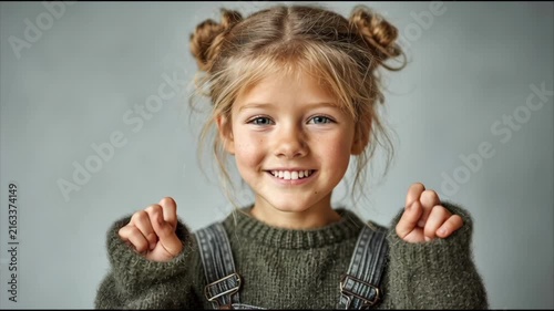 A happy young Caucasian girl wearing a green sweater and denim overalls making playful hand gestures against a minimalist gray studio background.
