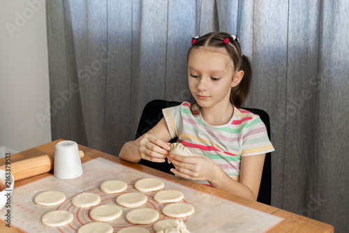 A little girl of european appearance sits at a wooden table, carefully pinching the edge of a homemade dumpling or pierogi during the cooking process. Home cooking concept.