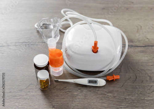 Close-up of a medical nebulizer with inhalation mask, digital thermometer, and medicine bottles on a wooden table. Concept of respiratory therapy, healthcare at home, and asthma treatment