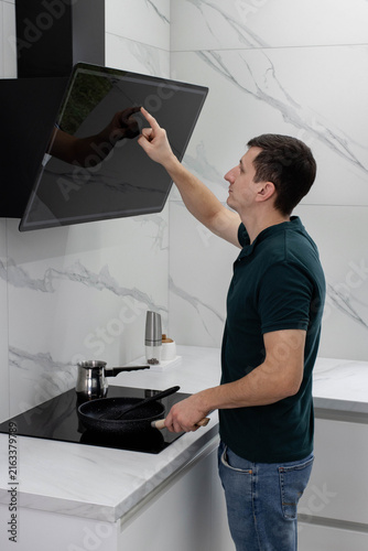A young man stands by the induction hob, holding a frying pan and touching the control panel of a modern black cooker hood. Concept of home life and daily household chores. Bachelor's life.