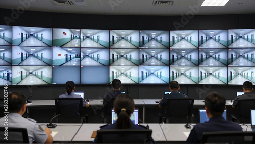 Prison security officers monitor corridor surveillance screens in a central control room